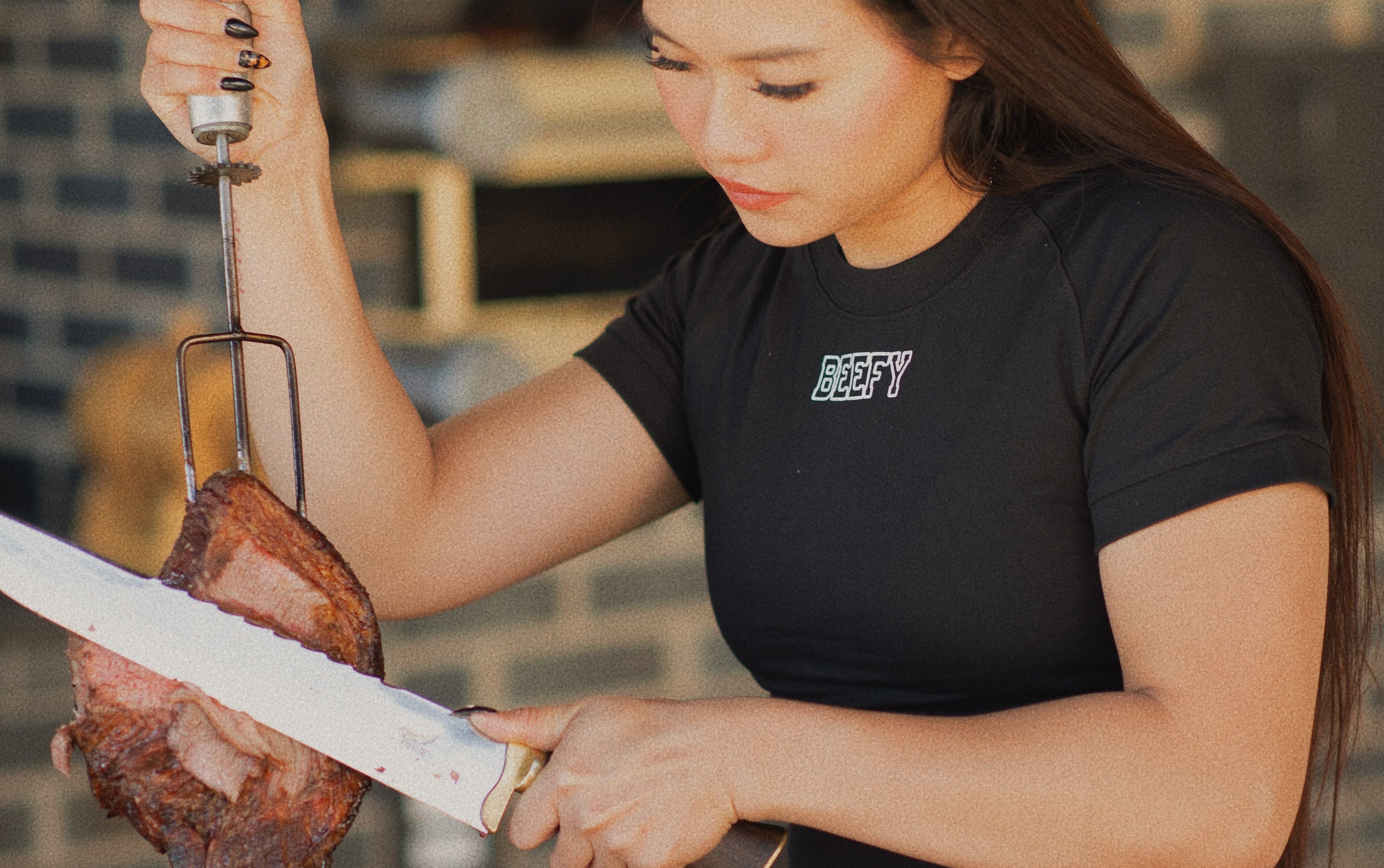 Woman preparing meat on a skewer outdoors with a brick wall background, wearing a shirt that says "beefy"