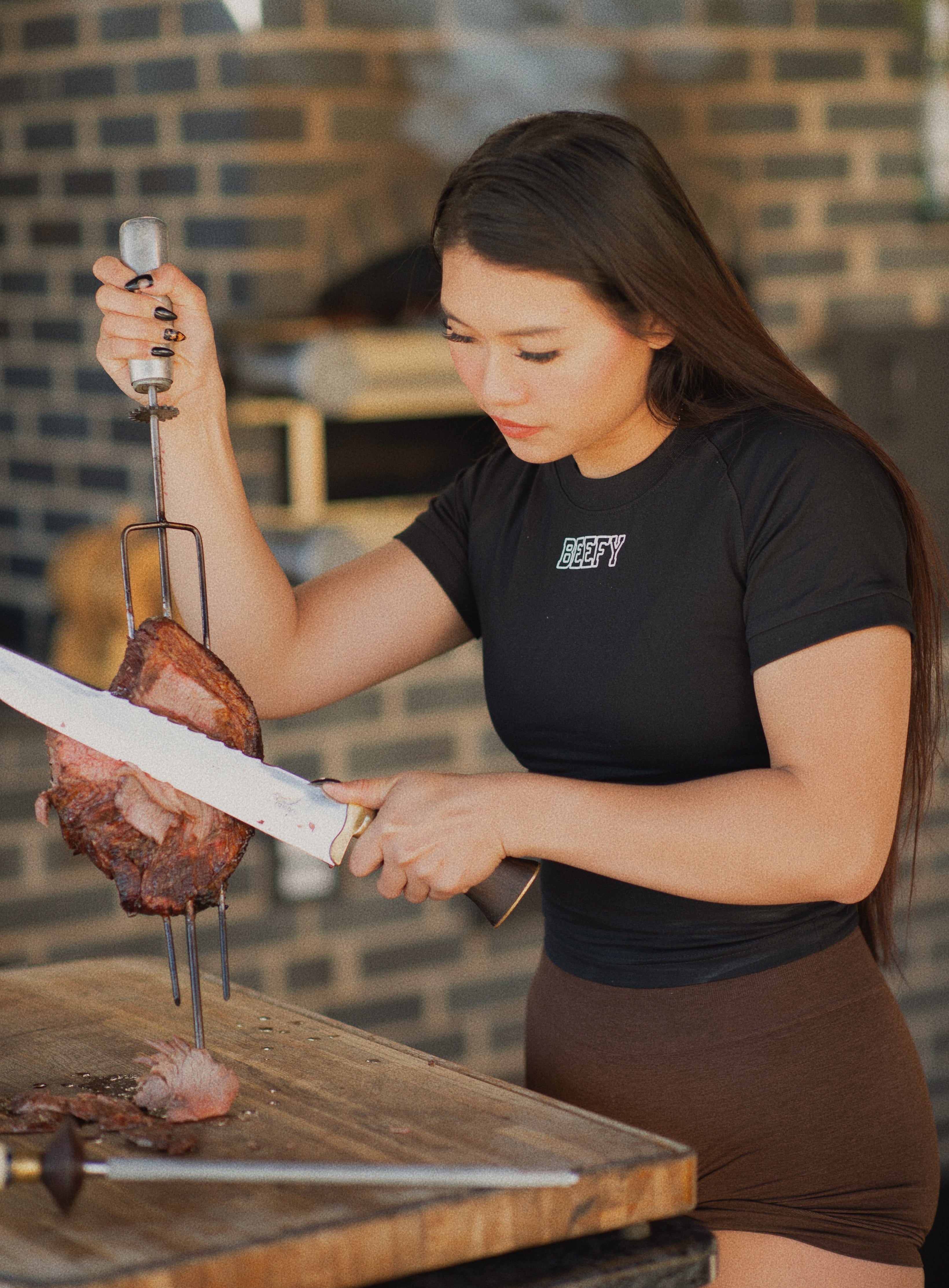 Woman preparing meat skewers with a brick wall background, wearing a shirt that says "beefy"