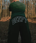 A person wearing green cargo sweats with the word 'BEFFY' printed on the back, standing in a wooded area.