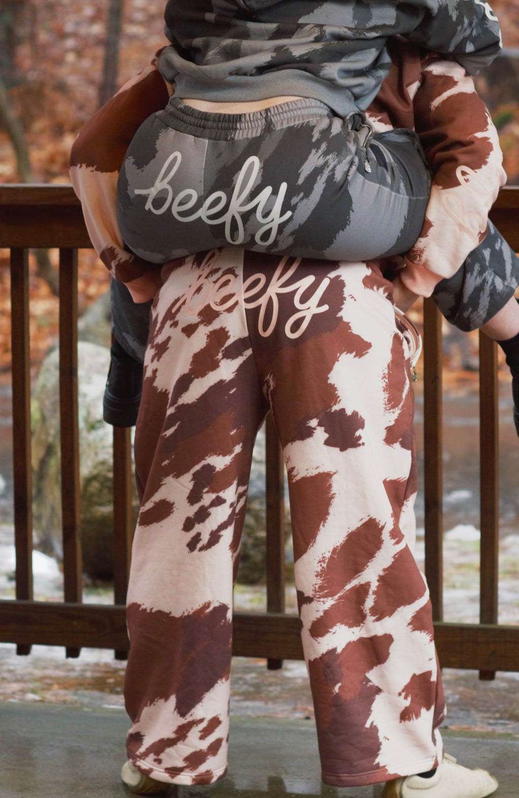 Two people wearing cow-print pants with "beefy" on them giving each other piggy back rides